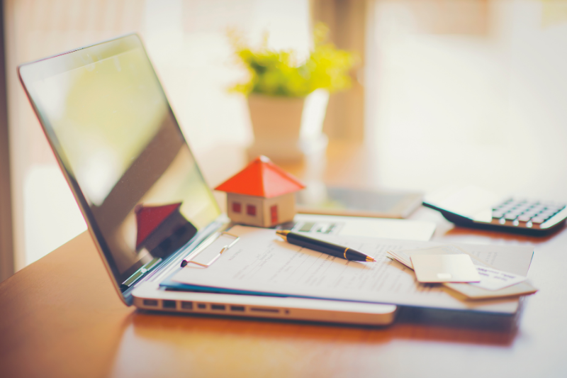 Home 121 A laptop, paperwork, a pen, credit cards, a small model house, and a calculator are arranged on a desk with a potted plant in the background, suggesting a work-from-home or real estate theme.