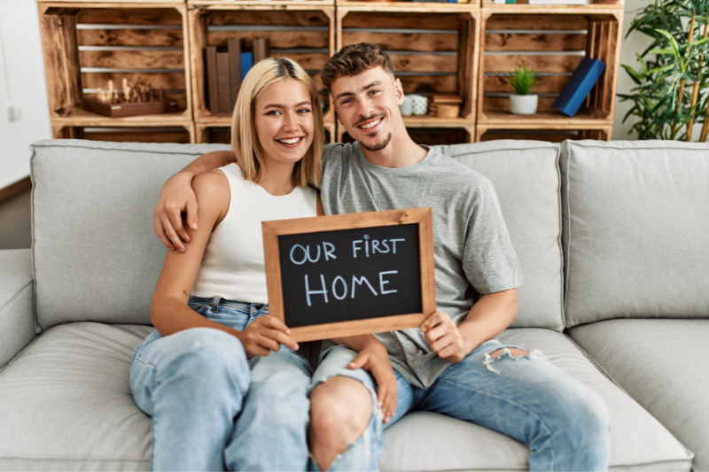 Home 115 A smiling couple sits on a couch holding a chalkboard sign that reads “Our First Home,” with wooden shelves and plants in the background.