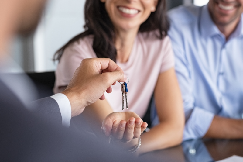 Home 114 A person hands a set of keys to a smiling woman, who is reaching out her hand, while another person sits beside her. The scene suggests the purchase or rental of a new home.