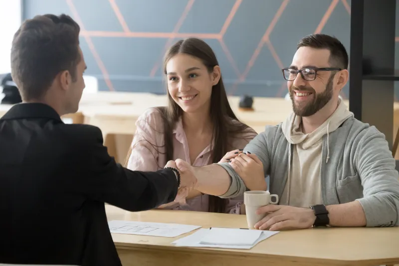 Home 111 A man and woman smile while sitting at a desk, shaking hands with another man across from them. Papers and a coffee cup are on the table, suggesting a successful meeting or agreement.