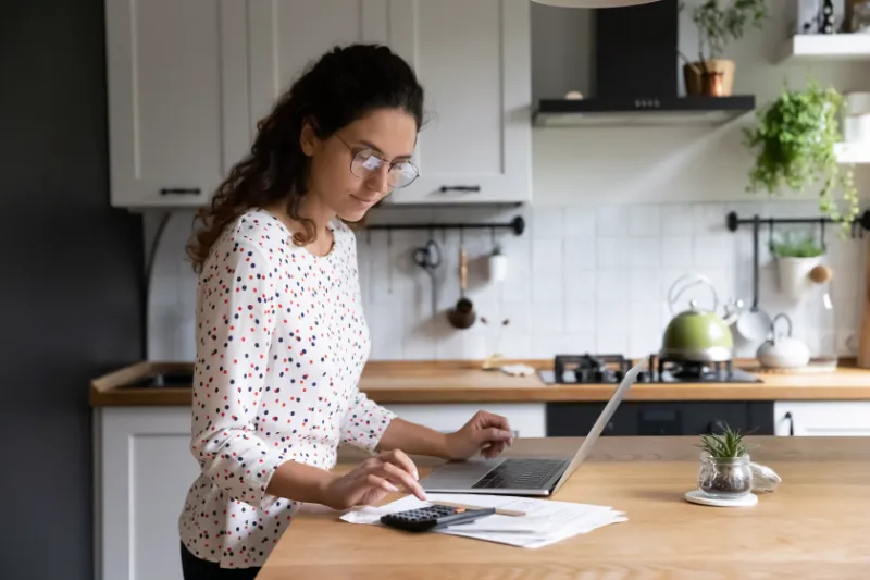 Home 110 A woman stands in a kitchen, using a laptop and a calculator, with papers spread out on the counter. She appears focused, wearing glasses and a polka-dot shirt. Plants and kitchenware are visible in the background.