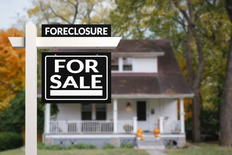 Home 106 A white post with Foreclosure and For Sale signs stands in front of a white house with a porch, surrounded by trees with autumn leaves. The house appears to be vacant.
