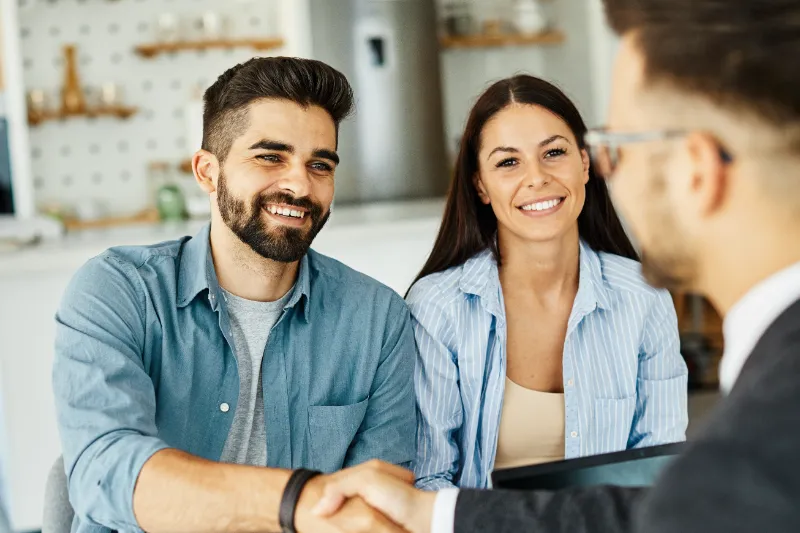 Home 109 A smiling man and woman sit together at a table, shaking hands with another person in business attire across from them in a bright, modern office setting.