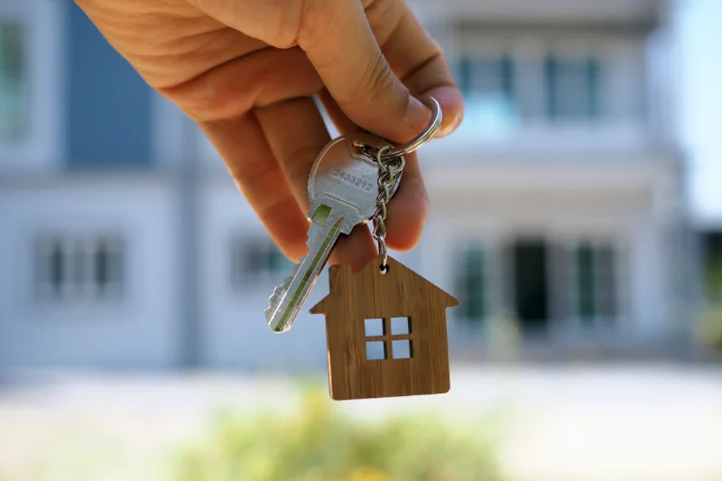 Home 105 A hand holding a house key and a keychain shaped like a small house, with a blurred building in the background.
