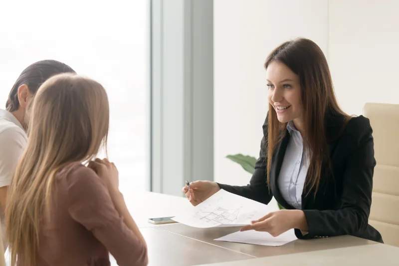 Home 102 A smiling woman in a business suit sits at a desk, holding papers and a pen, talking to two people seated across from her in a bright office setting.