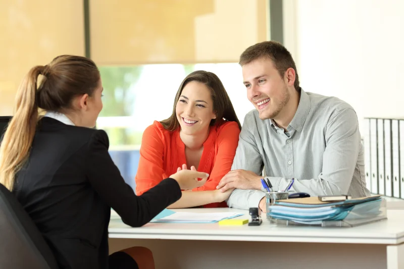 Home 101 A smiling couple sits at a desk across from a businesswoman, engaged in a friendly conversation. Papers, a pen, and eyeglasses are on the desk in a bright, modern office setting.