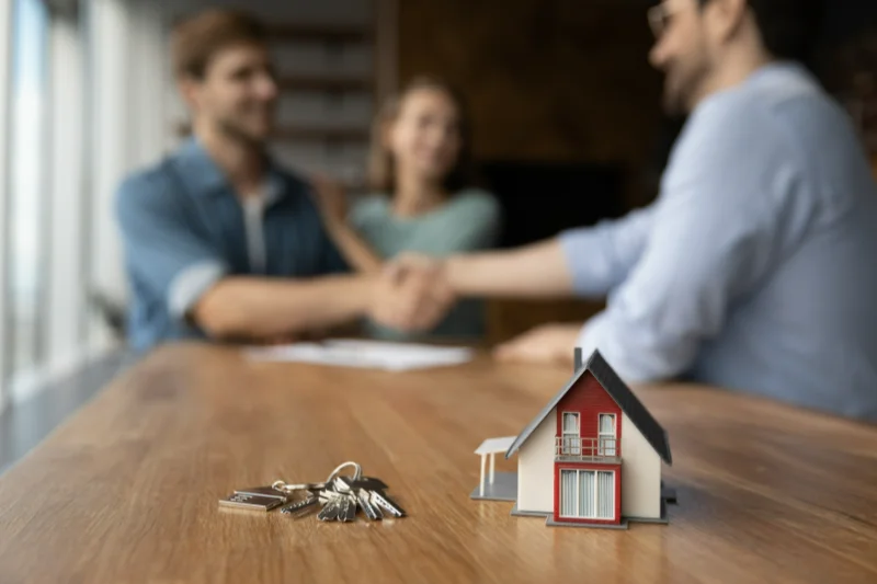 Home 103 A miniature house and keys sit on a wooden table in focus, while two people shake hands and a third person looks on in the blurred background, suggesting a real estate transaction.