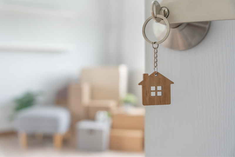 Home 97 A house-shaped keychain hangs from a key in a door, with packed boxes and furniture visible in a softly focused room, suggesting moving into a new home.