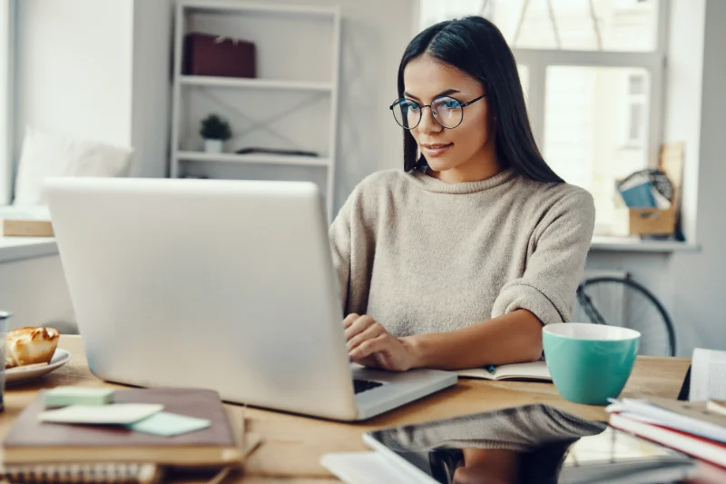 Home 100 A woman with long dark hair and glasses is sitting at a desk, working on a laptop. She is surrounded by notebooks, a mug, and office supplies in a bright, modern workspace.