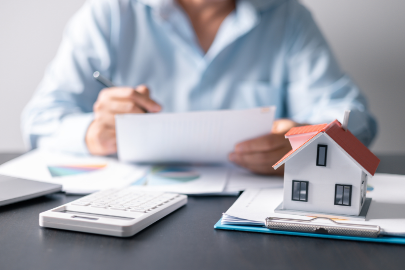 Home 96 A person sits at a desk reviewing documents with charts, next to a calculator and a model house, suggesting work related to real estate or financial planning.