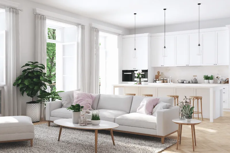 Home 93 A bright, modern open-plan living room and kitchen with white furniture, light wood accents, potted plants, large windows, and pendant lights. Sunlight streams in, creating an airy, inviting atmosphere.