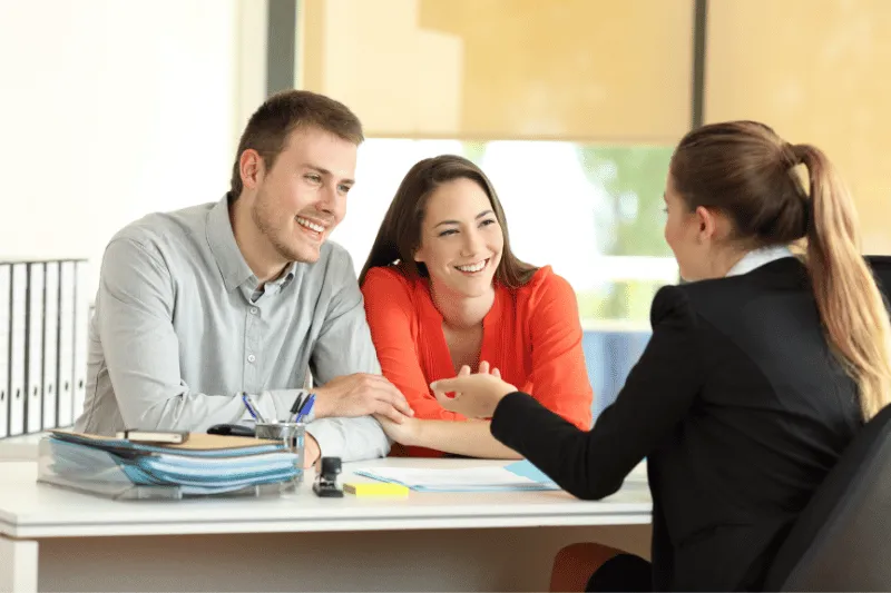 Home 94 A smiling couple sits at a desk across from a professional woman in an office, having a friendly discussion. The couple appears attentive and engaged, with documents and folders on the desk.