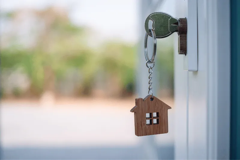 Home 95 A silver key with a wooden house-shaped keychain is inserted into a door lock, with an outdoor background softly blurred.