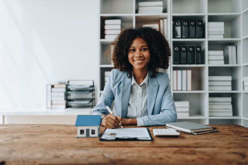 Home 89 A woman in a light blue blazer sits at a desk, smiling, with documents, a pen, a small house model, a notebook, and a calculator in front of her. Bookshelves with files and books are in the background.