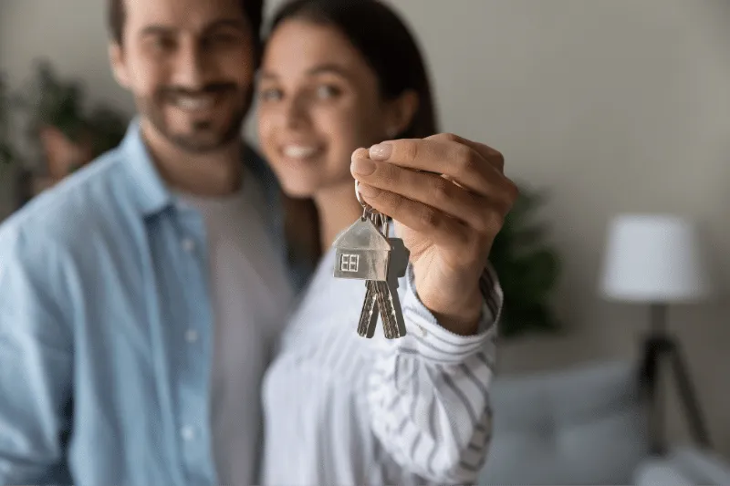 Home 91 A smiling couple stands close together, with the woman holding out a set of house keys toward the camera, suggesting they have just bought a new home. The background is softly blurred indoors.