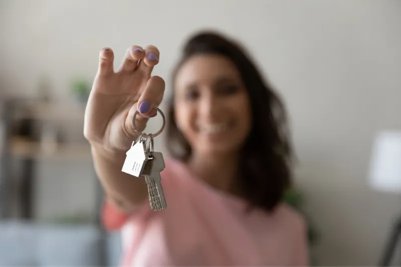 Home 87 A smiling woman holds a set of keys with a house-shaped keychain toward the camera, suggesting new home ownership. The background is softly blurred.
