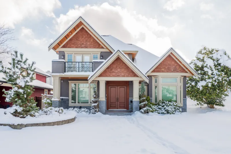 Home 86 A two-story house with a peaked roof and large front windows is surrounded by snow-covered trees and yard under a bright, partly cloudy sky.