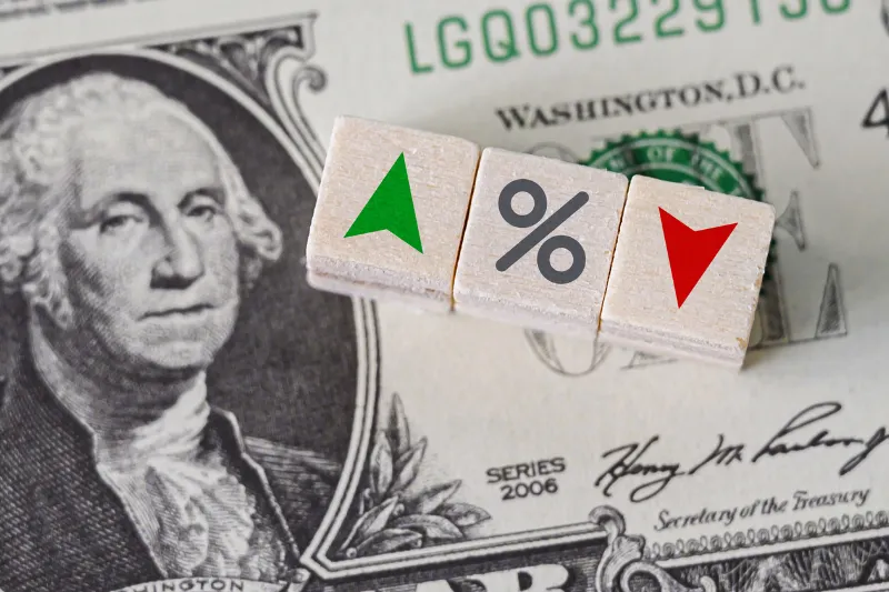 Home 85 A close-up of a U.S. one-dollar bill with three wooden blocks on top displaying a green upward arrow, a percentage symbol, and a red downward arrow.