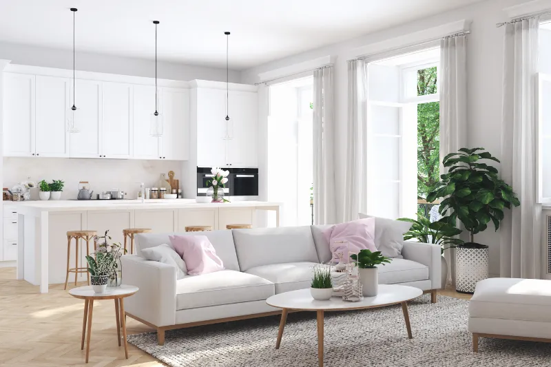 Home 83 A bright, modern living room with white sofas, light wood furniture, green plants, and large windows letting in natural light. An open kitchen with white cabinets and pendant lights is visible in the background.