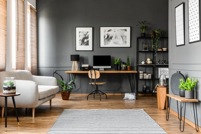 Home 82 Modern home office with dark gray walls, wooden desk with computer, chair, various plants, and minimalist decor. A beige sofa and a striped rug are in the foreground, shelves with books and decor items are on the right.