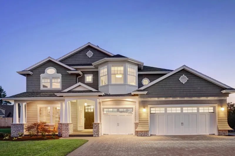 Home 84 Two-story suburban house at dusk with gray siding, white trim, large front porch, three-car garage, and illuminated windows. The driveway and small landscaped yard are visible in front.