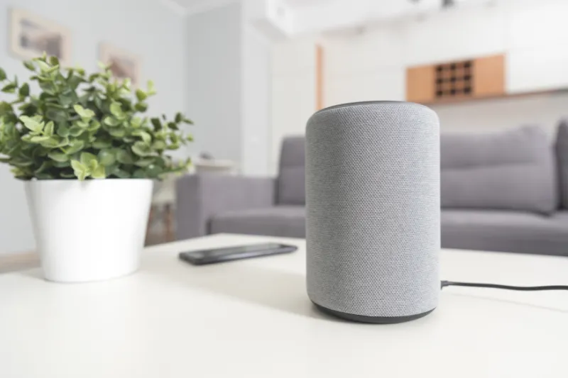 Home 81 A gray smart speaker sits on a white table next to a potted green plant and a smartphone, with a sofa and shelves visible in the blurred background of a modern living room.