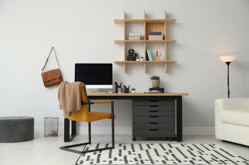 Home 80 A modern home office with a desk, computer, yellow chair, tan sweater, wall shelves holding books and decor, a brown bag hanging on the wall, a floor lamp, and a white sofa on a patterned rug.