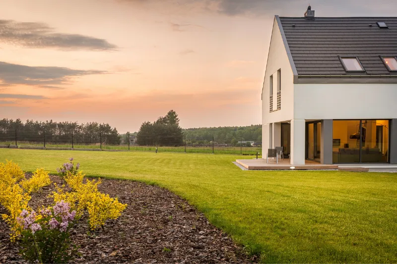 Home 78 Modern house with large windows and sloped roof, set on a spacious green lawn with yellow and purple flowers, under a cloudy sunset sky, with trees and a fence in the background.