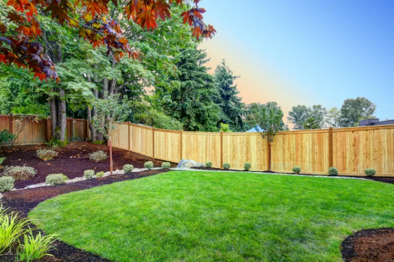 Home 76 A backyard with green grass, small shrubs along the edges, mulched flower beds, and a wooden privacy fence. Tall trees and clear sky are visible in the background.