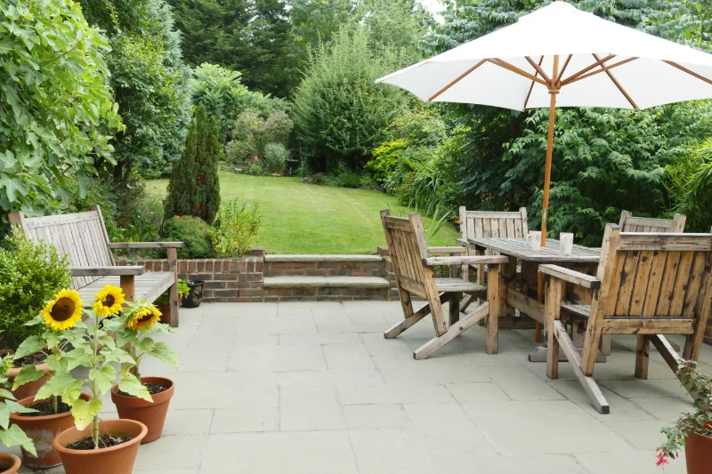 Home 75 A patio with wooden benches and a dining table set beneath a large white umbrella, surrounded by potted sunflowers and lush green garden foliage in the background.
