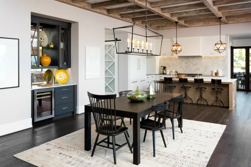 Home 73 Modern open-concept kitchen and dining area with dark wood floors, black dining table and chairs, white cabinets, bar stools at a kitchen island, pendant lighting, and exposed wood-beam ceiling.