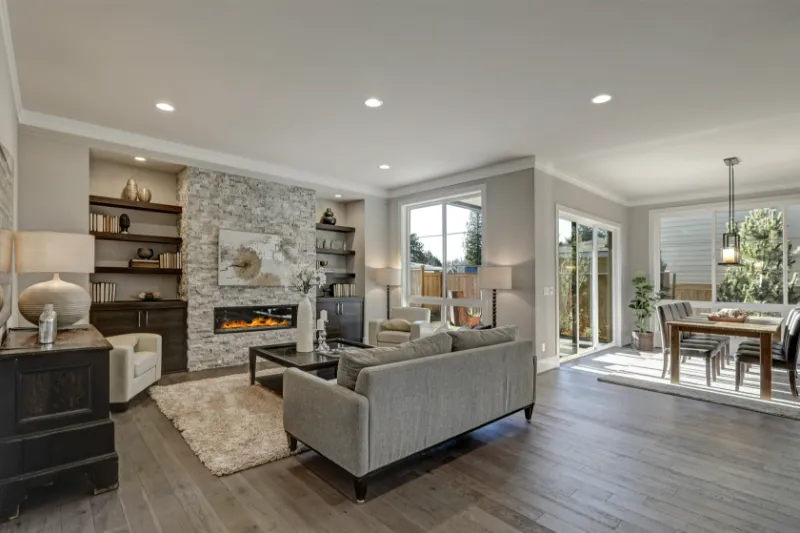 Home 74 Modern living and dining area with gray sofas, stone fireplace, built-in shelves, large windows, hardwood floors, and a dining table near sliding glass doors leading to a fenced backyard.