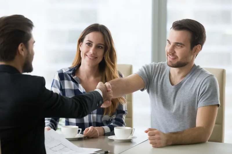 Home 72 A man and woman sit at a table with coffee cups, smiling as the man shakes hands with another man in formal attire, suggesting a successful meeting or agreement.