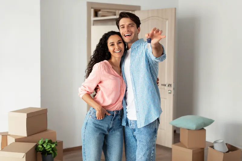 Home 71 A smiling couple stands close together in a room with moving boxes. The woman wears a pink shirt and jeans, and the man wears a blue checkered shirt and jeans, gesturing excitedly as they look toward the camera.