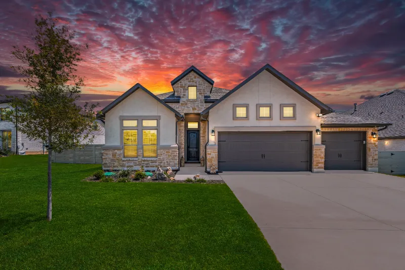 Home 67 A modern, single-story house with stone accents and a three-car garage is shown at sunset. The sky is vibrant with red and purple clouds, and the front lawn is neatly landscaped with a small tree and flower beds.