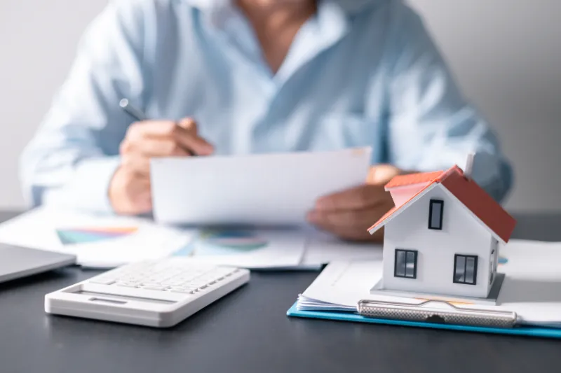 Home 65 A person in a blue shirt reviews documents at a desk with a calculator, paperwork, pen, and a small model house, suggesting real estate, mortgage, or financial planning.