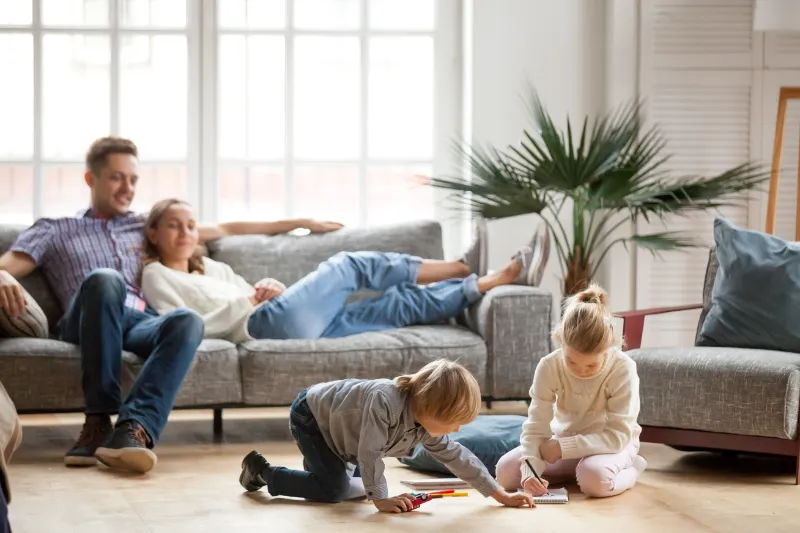 Home 66 A man and woman relax together on a sofa while two young children draw and color on the floor in a bright, cozy living room with large windows and a potted plant.