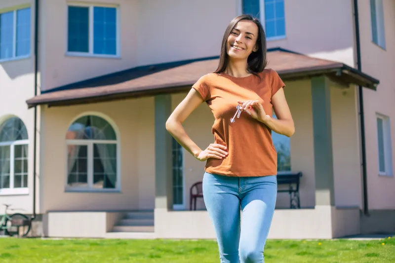 Home 62 A smiling woman stands in front of a house, holding a set of keys in her hand, suggesting she is a new homeowner. She is wearing a rust-colored T-shirt and blue jeans.