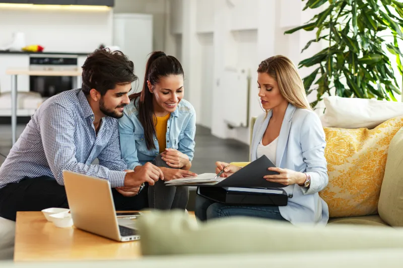 Home 63 A professional woman shows documents to a smiling couple seated on a sofa in a modern, bright living room. A laptop and coffee cup are on the table, and a large plant is in the background.