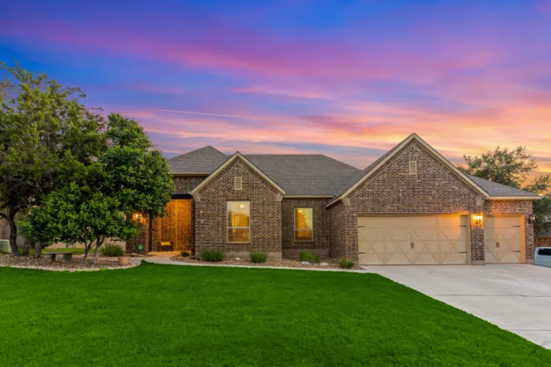 Home 64 Single-story brick house with a three-car garage, lit windows, and a well-kept green lawn, set against a colorful sunset sky with pink and purple hues. Trees and shrubs line the front walkway.