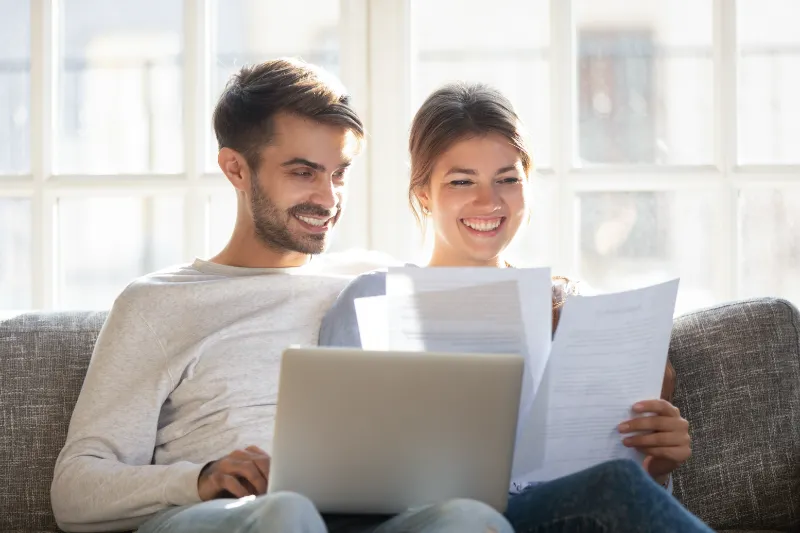 Home 60 A smiling couple sits on a couch, looking at documents together. The man has a laptop on his lap, and both appear happy and relaxed, sitting in a bright room with large windows in the background.