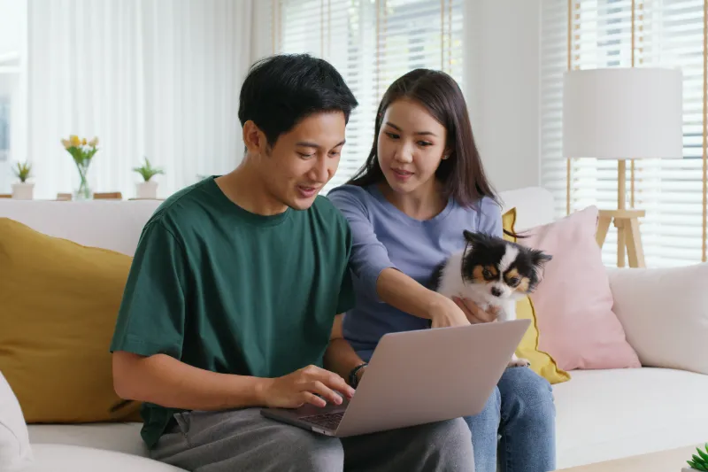 Home 59 A man using a laptop sits on a sofa next to a woman holding a small dog. The woman points at the laptop screen. They appear to be discussing something together in a bright, modern living room.