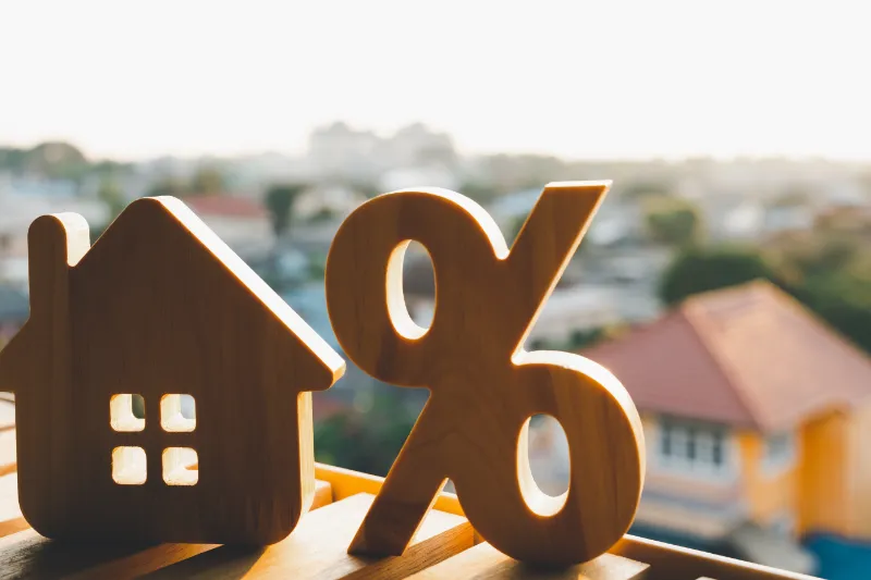 Home 58 Wooden house and percentage symbol figures on a table with blurred houses and sunlight in the background, representing mortgage or interest rates in real estate.