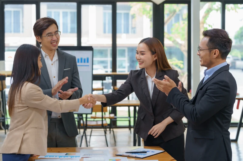 Home 56 Four business professionals in formal attire are in a bright office. Two women in the center are shaking hands, while two men beside them are smiling and clapping, suggesting a successful meeting or agreement.