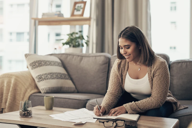 Home 55 A woman sits on a sofa in a cozy living room, smiling as she writes in a notebook on a coffee table. Papers, a mug, and glasses are also on the table. Shelves and windows are visible in the background.