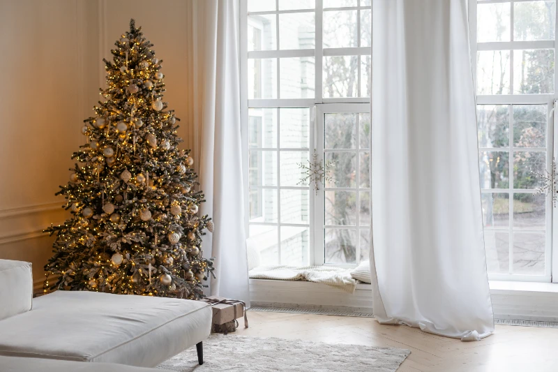 Home 53 A decorated Christmas tree with white and gold ornaments and lights stands in a bright living room next to large windows with white curtains. A few wrapped gifts sit beneath the tree.