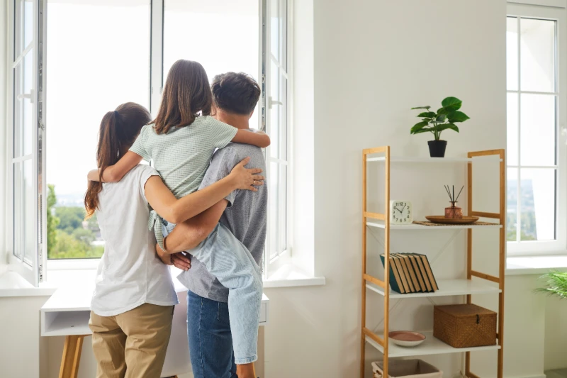 Home 49 A family of three, seen from behind, stands by an open window. The father holds their child, while the mother stands close. Sunlight fills the bright, tidy room with shelves, a clock, boxes, and a potted plant.