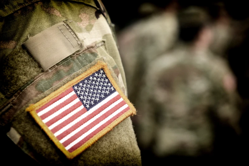 Home 47 Close-up of a U.S. flag patch on the sleeve of a camouflage military uniform, with other blurred soldiers in similar uniforms in the background.