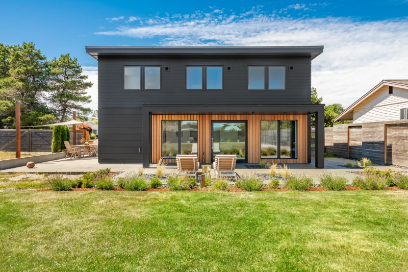 Home 46 Modern two-story house with dark siding and large windows, wooden accents around the doors, a patio with two lounge chairs, and a landscaped yard with green grass and garden beds under a blue sky.