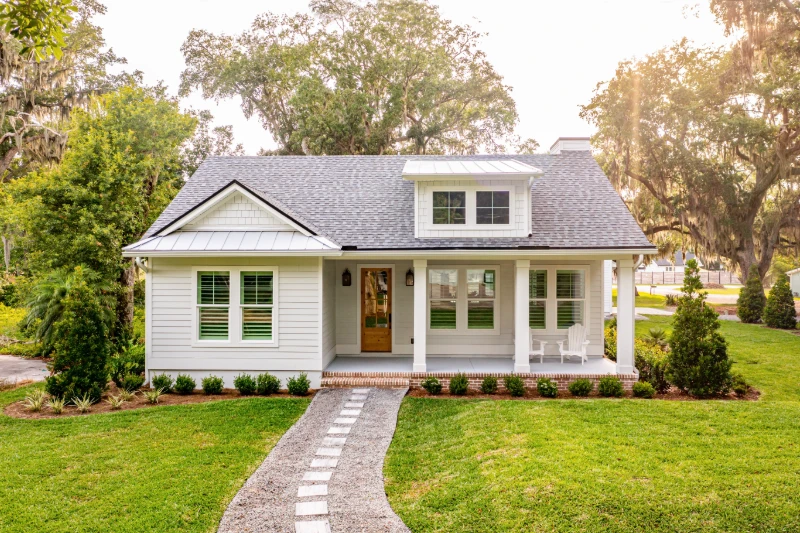 Home 48 A charming white cottage with a front porch, brick steps, and two rocking chairs, surrounded by green grass, shrubs, and tall trees on a sunny day.